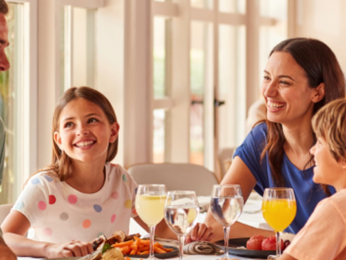 A family with two children laughing over a meal at a table by tall sunlit windows.