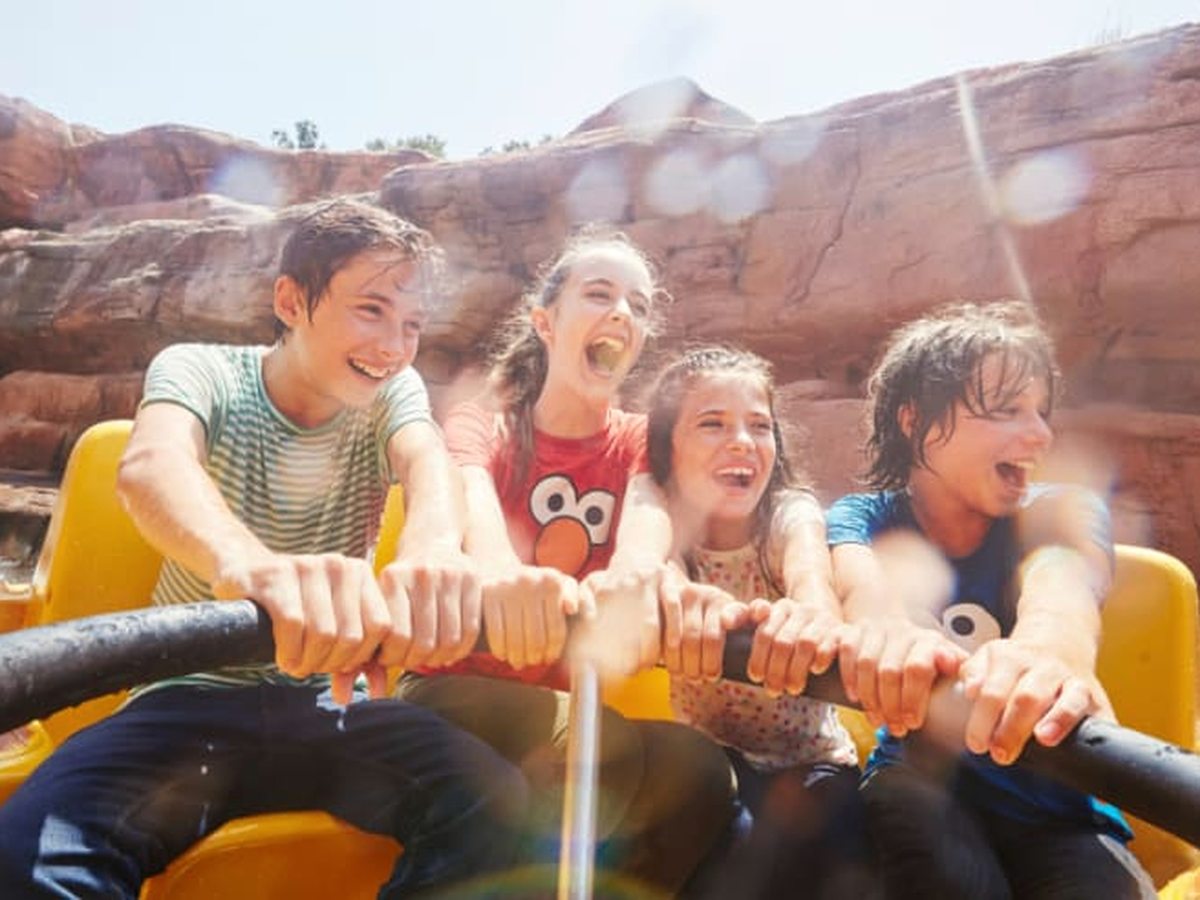 Four children gripping the handrail of a yellow water-flume boat, laughing as it splashes through a red-rock canyon.