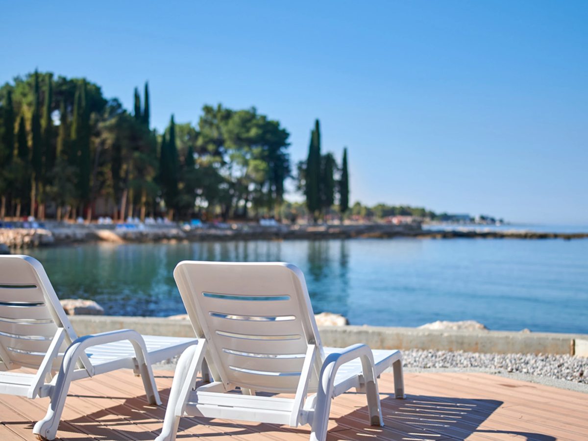 Two white sun loungers on a paved timber deck at the water's edge, looking across a calm bay to a wooded point.