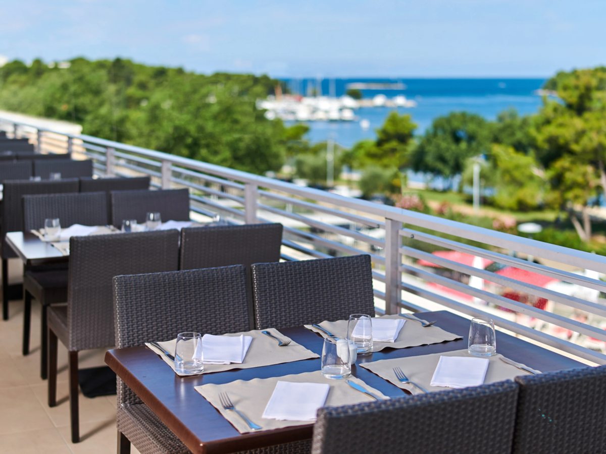 Dark woven chairs and tables laid for a meal on an open terrace, with a marina and the sea beyond the railing.
