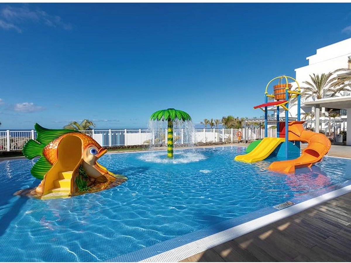 A shallow children's pool with a fish tunnel, a palm-shaped water sprayer and small slides, fenced, with the tower behind.