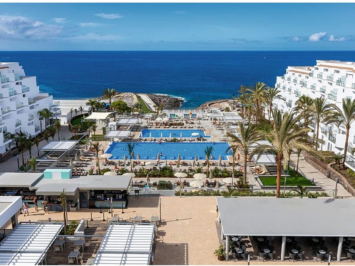 Aerial view of Hotel Riu Buenavista, two white accommodation wings set around large blue pools on the coast, open sea beyond.