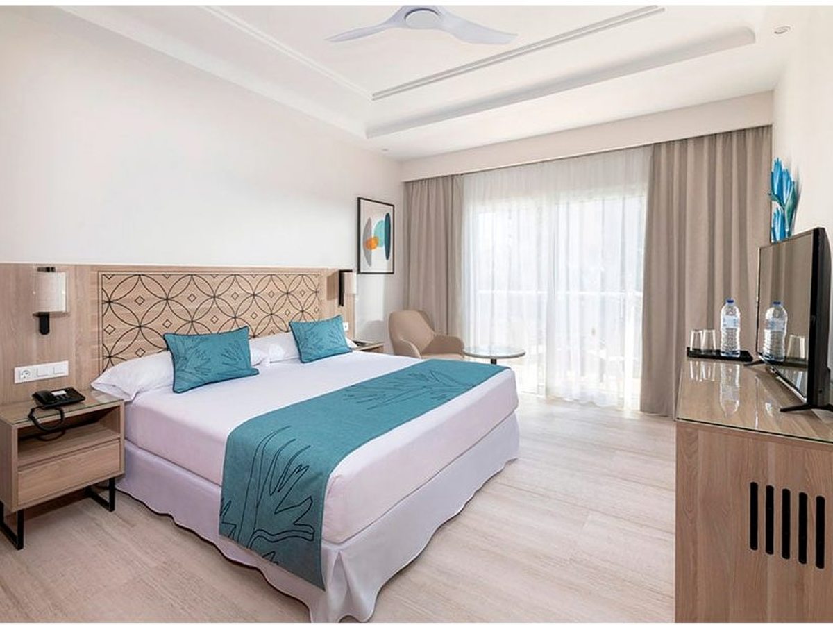 Double room with a patterned wooden headboard, a teal bed runner and cushions, a lounge chair and balcony doors letting in daylight.