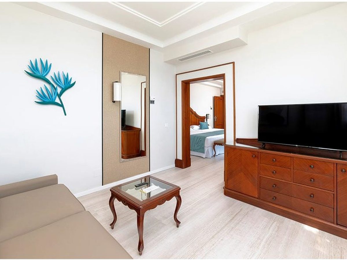 Suite sitting area with a sofa, a glass coffee table and a wood console below a TV, with an open doorway through to the bedroom.