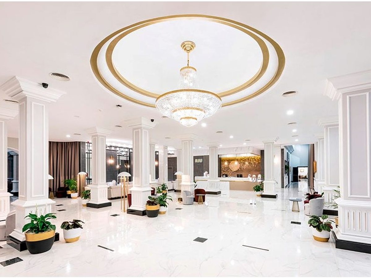 Bright white-marble lobby with white columns, a gold ceiling ring and chandelier, and the RIU reception desk beyond.