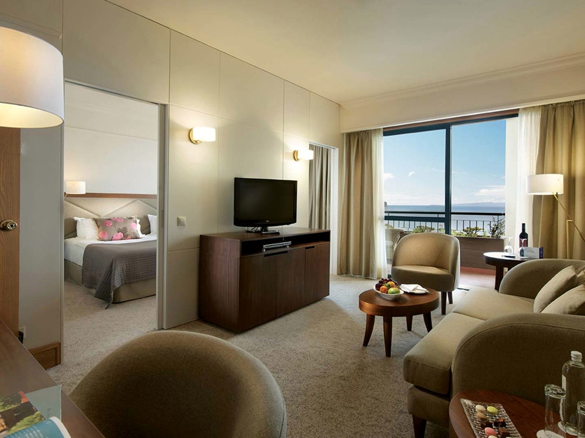 A suite sitting area with sofas, armchairs and a TV, the bedroom through a wide opening and a balcony with a sea view.