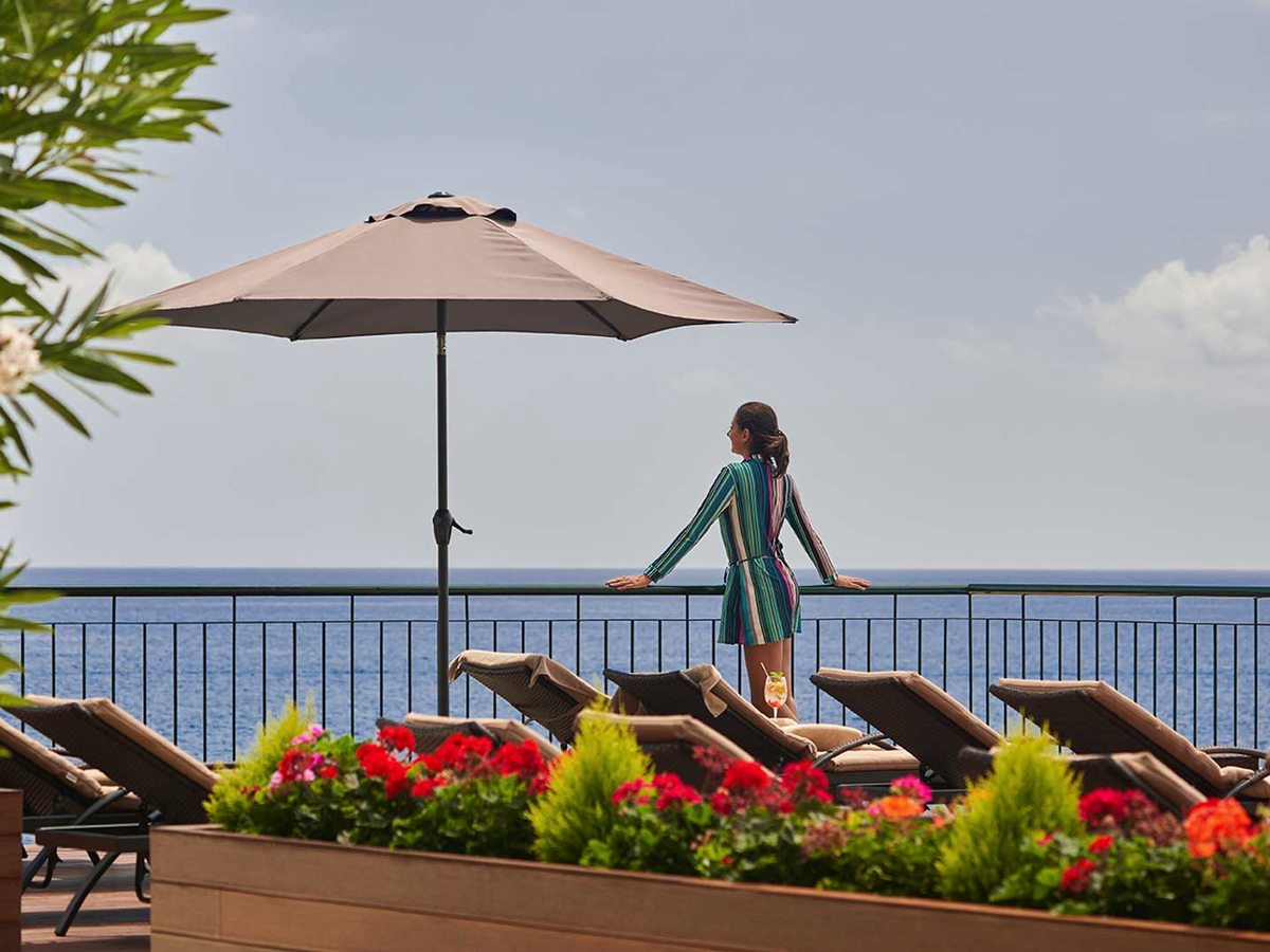 A sun terrace with cushioned loungers and planters of red geraniums under a parasol, a guest at the railing looking out over the open sea.