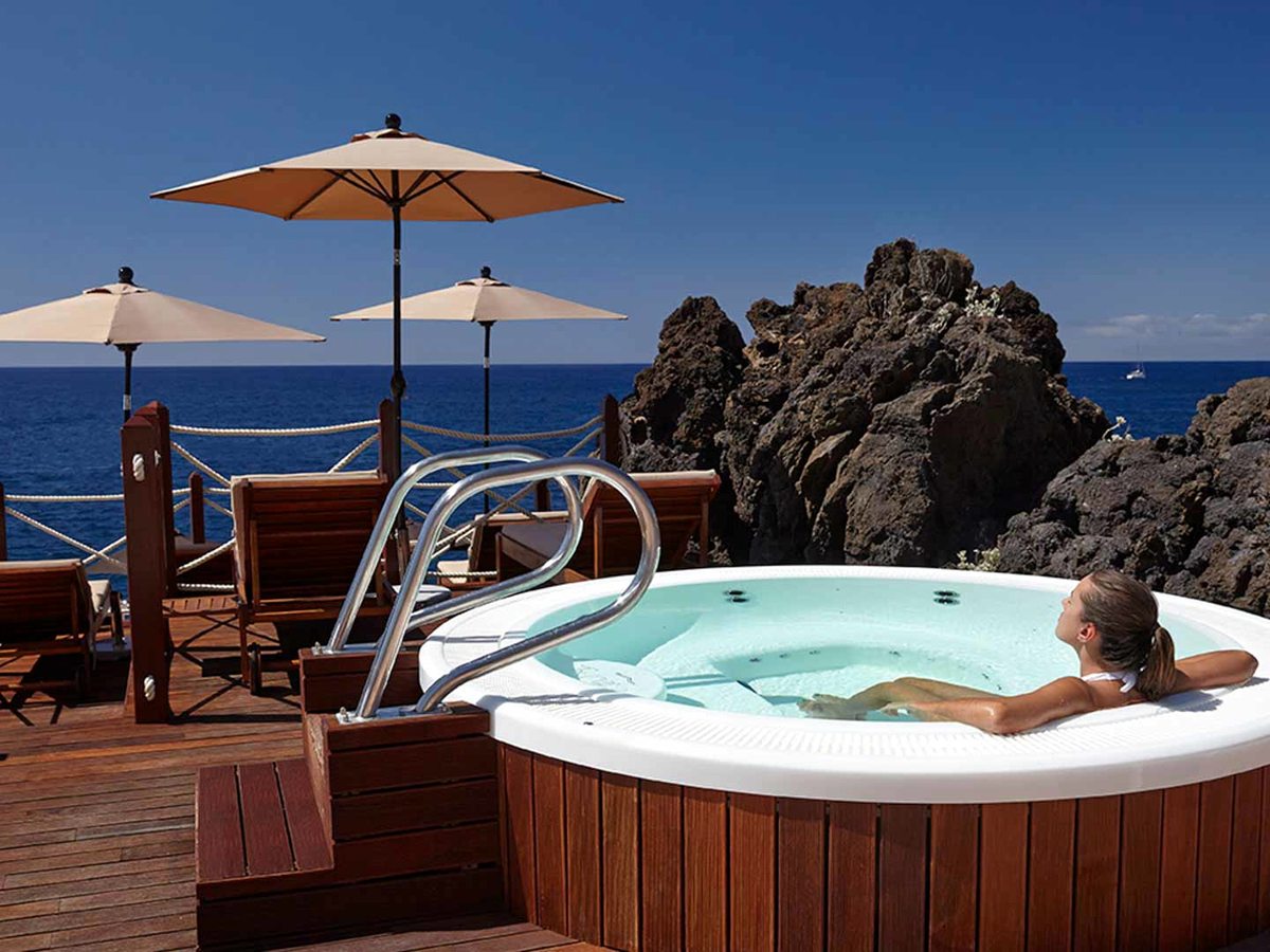 A round wooden hot tub on a timber deck with a bather in it, parasols and the sea behind.