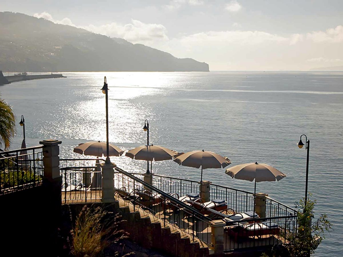 A stepped sea-view terrace with parasols and loungers above the water, the far headland of the Funchal coastline in the afternoon haze.
