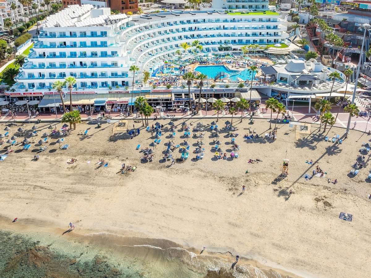 An aerial view of a golden-sand beach lined with straw parasols below the hotel and promenade shops.