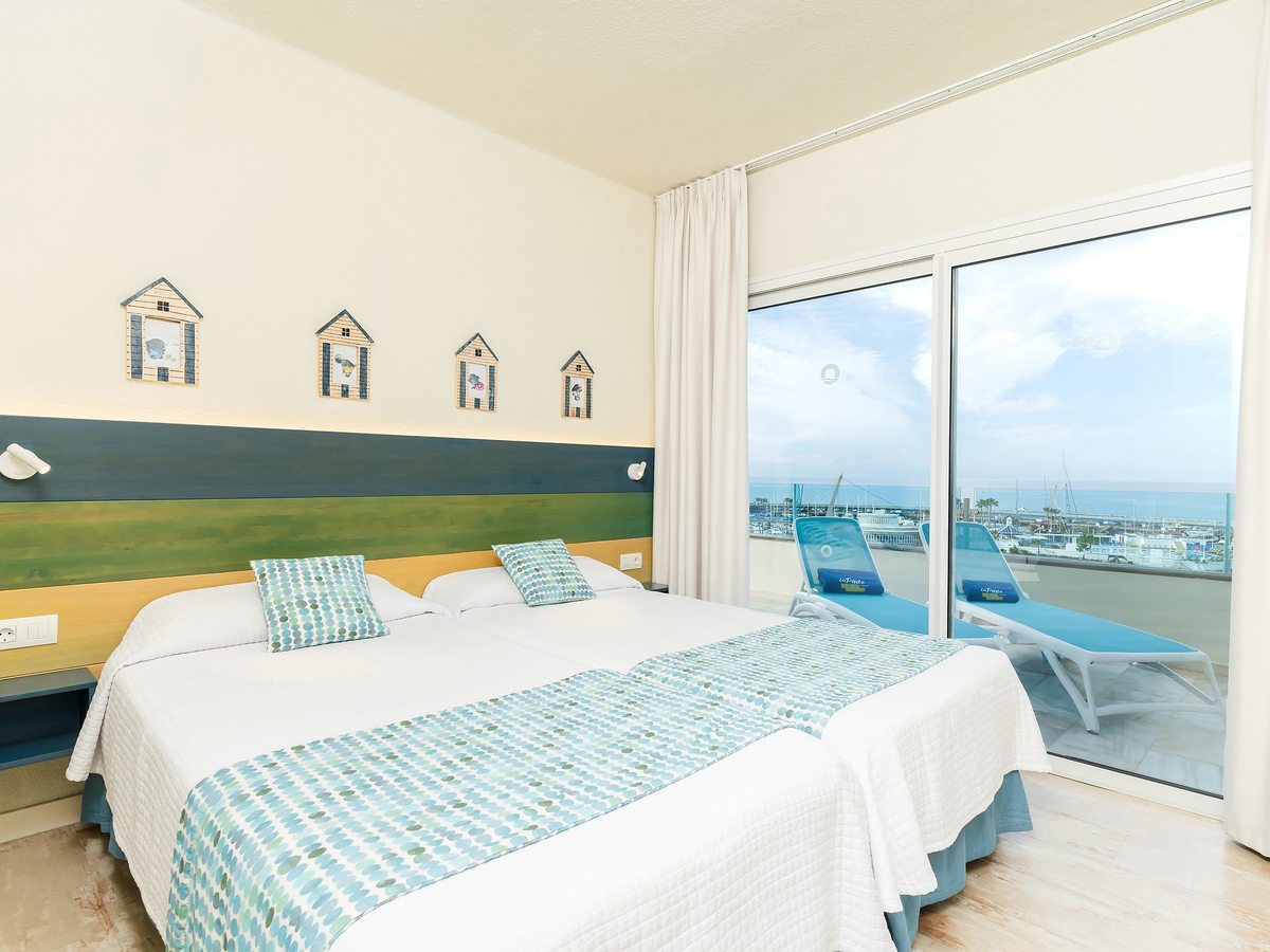A twin bedroom with glass doors to a balcony with a lounger and a marina view.