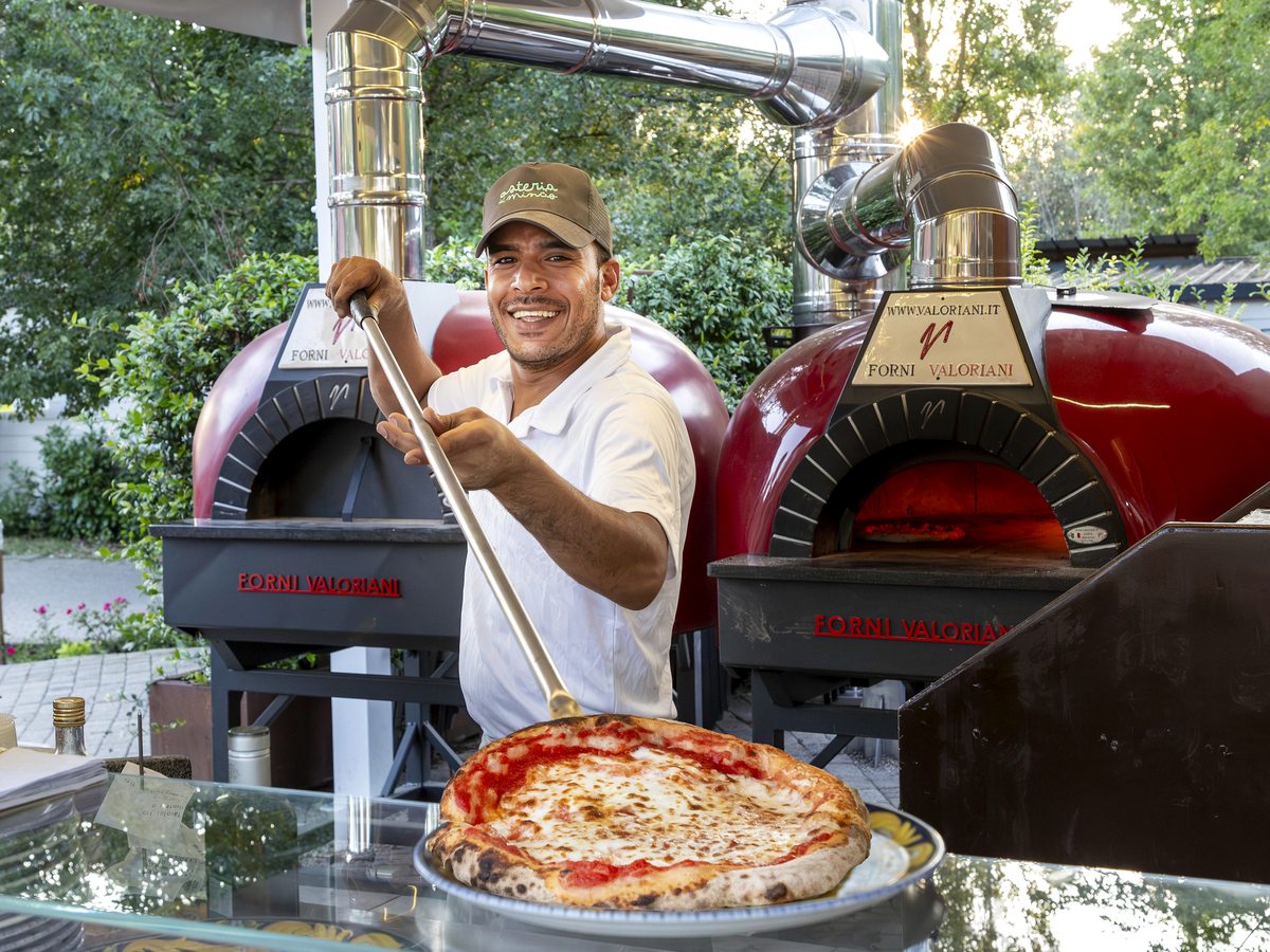 A chef sliding a margherita pizza from a red wood-fired oven on a long peel.