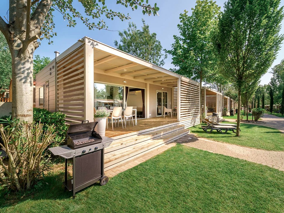 A modern mobile home with a covered terrace and a gas barbecue on the lawn beside it.
