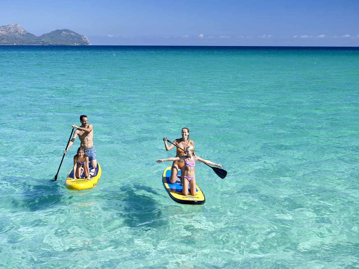 A family of four stand-up paddleboarding on two boards on calm clear turquoise water off the beach, mountains in the distance.