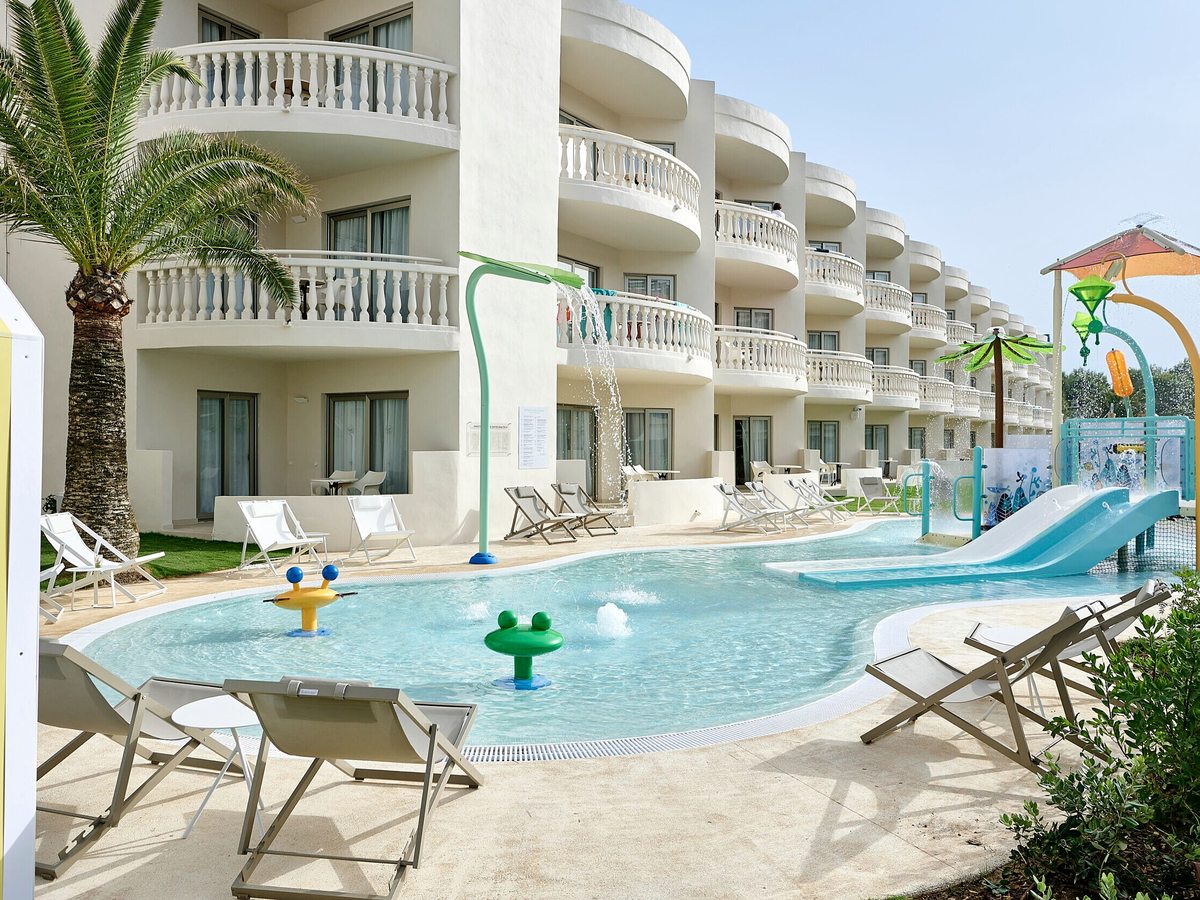 A shallow children's pool with a small blue water slide, sunloungers and ground-floor room balconies behind.