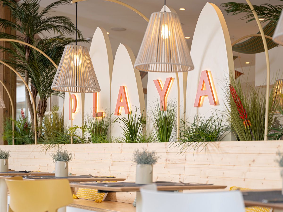 An indoor restaurant with set tables, pale yellow and white chairs, woven pendant lamps and the word PLAYA lit up in coral letters against surfboard-shaped panels and planting.