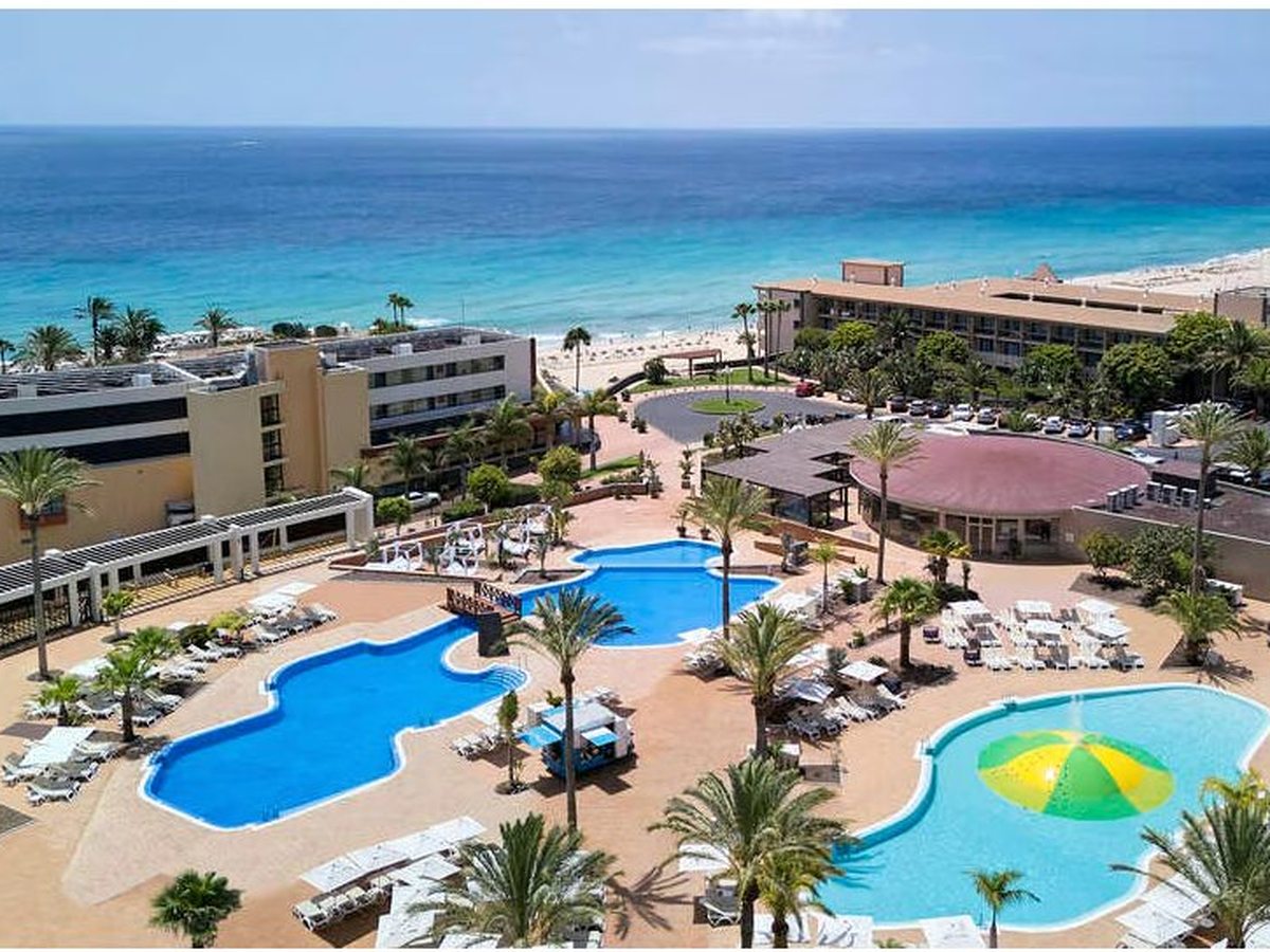 An aerial view of a cluster of freeform pools ringed by sun loungers and palm trees, with the hotel buildings and the golden beach and blue sea behind.