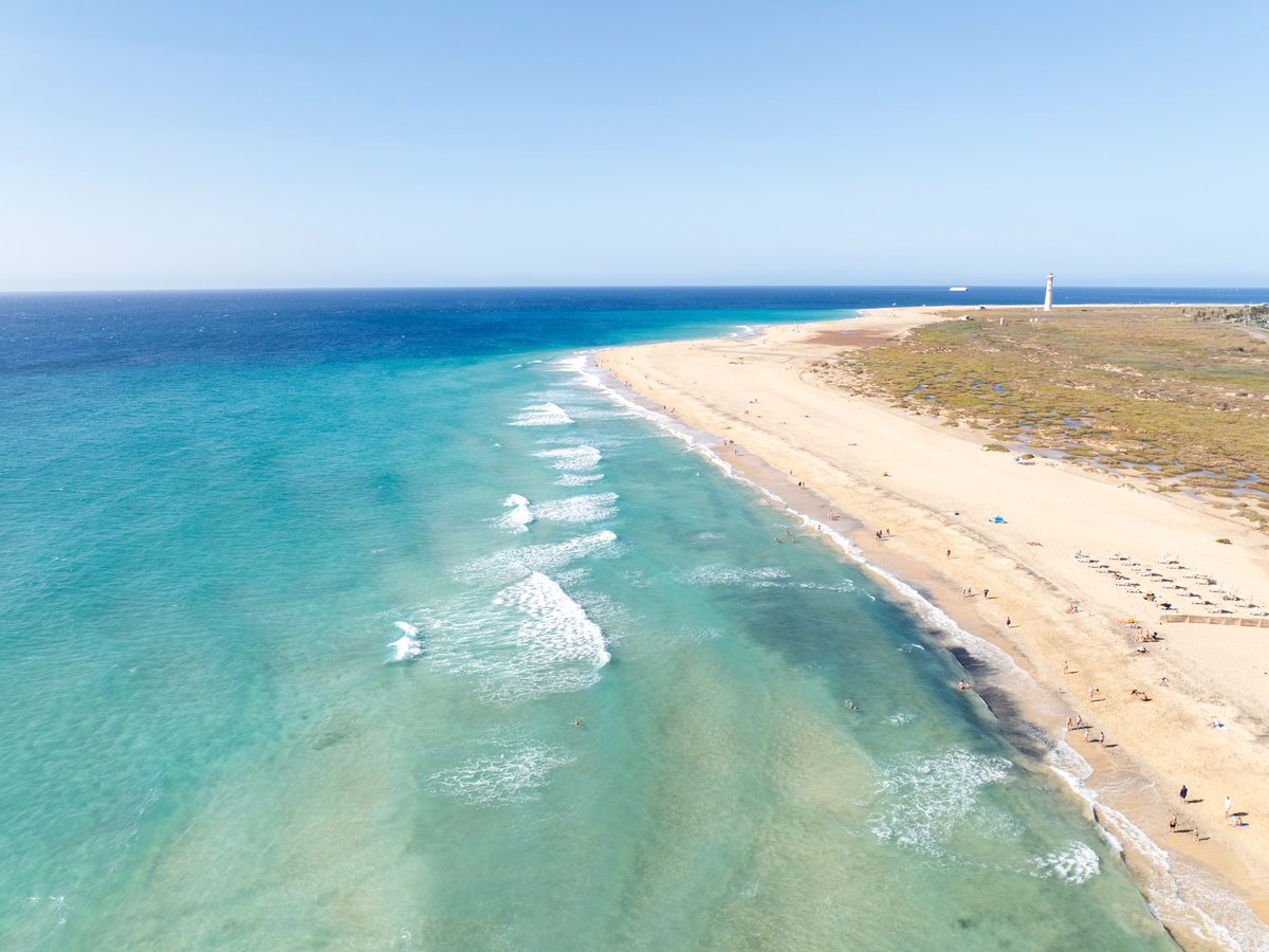 An aerial view along a wide golden-sand beach meeting turquoise shallows, with gentle waves and a lighthouse in the far distance.