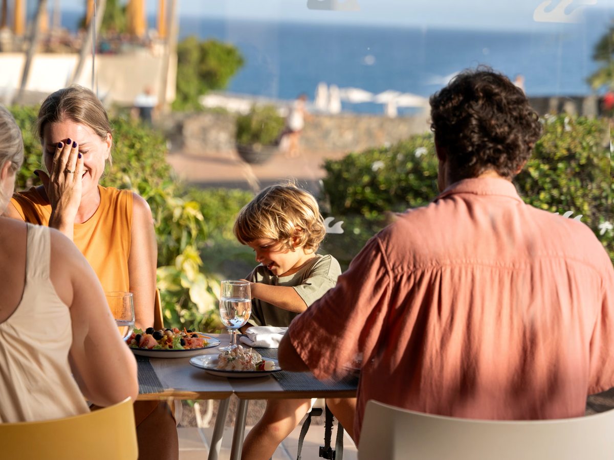 A family sitting at an outdoor table with plated food and drinks, greenery around them and the sea in the distance.