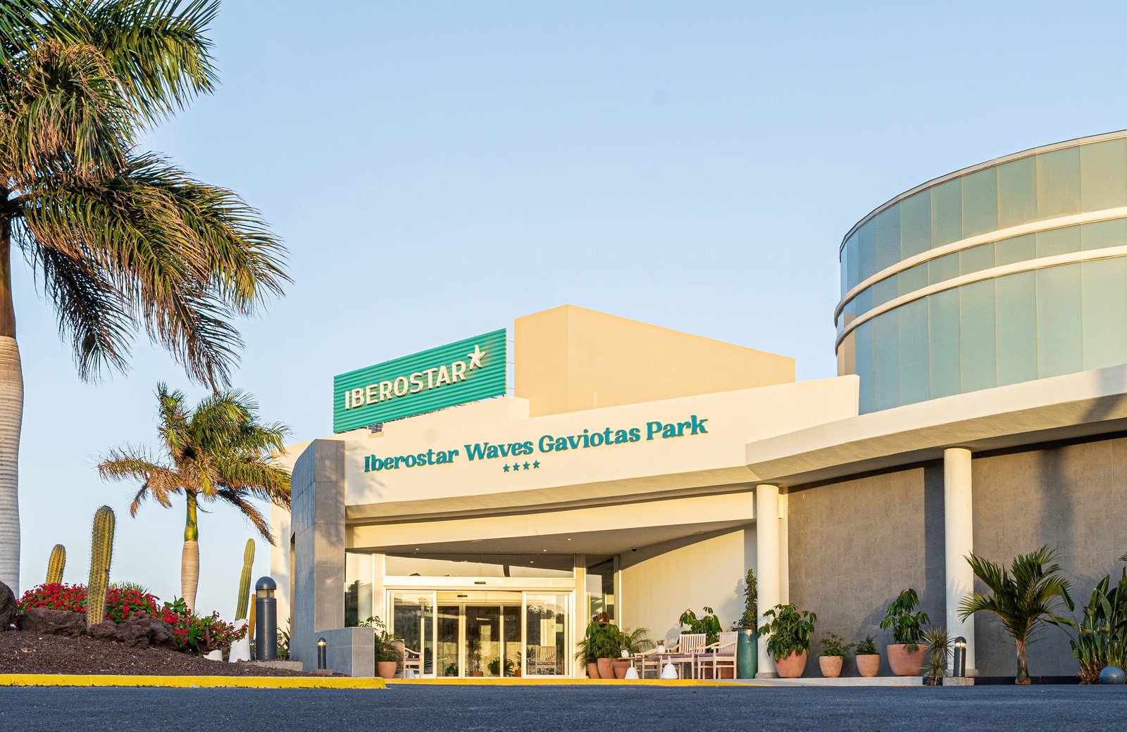 The main entrance to Iberostar Waves Gaviotas Park, with the resort's name above the doors, palms and cactus planting to the side and a clear blue sky.