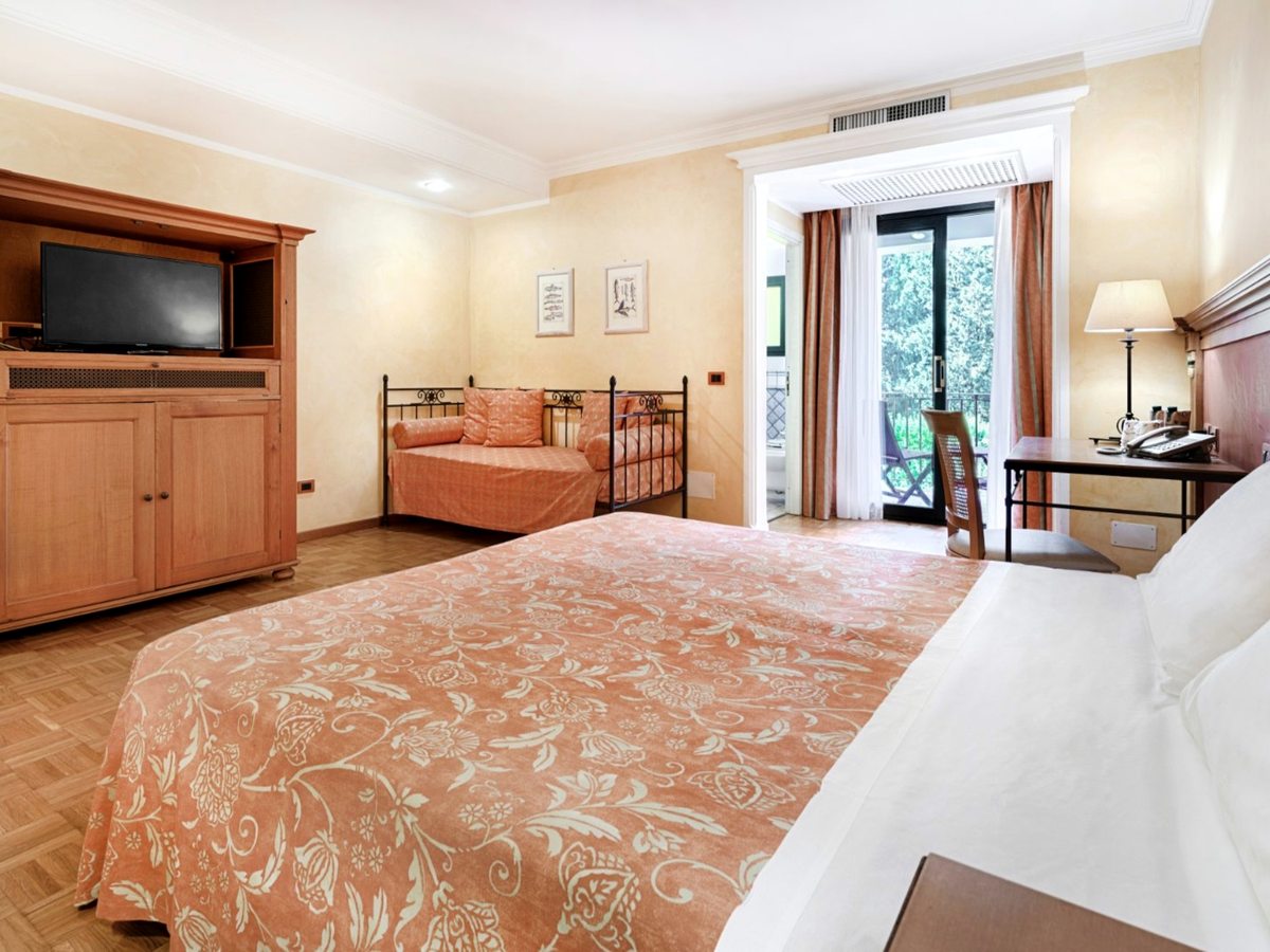 A double room with a light bedspread, a wooden TV cabinet and a writing desk, glazed doors open to a garden terrace.