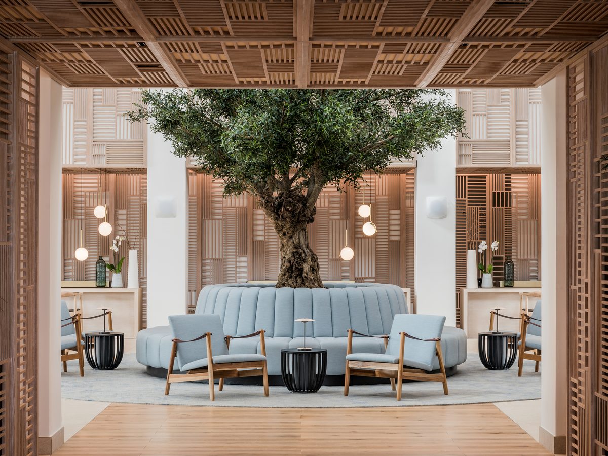 The reception lobby at Ikos Andalusia, a circular pale blue sofa set around an olive tree under a slatted timber ceiling.
