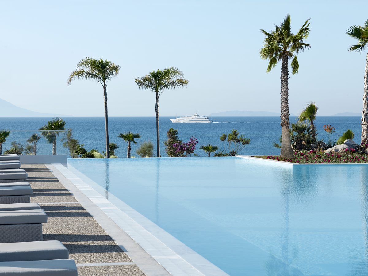 The main pool running to an infinity edge, with sun loungers along the tiled surround, palms behind and the sea beyond.