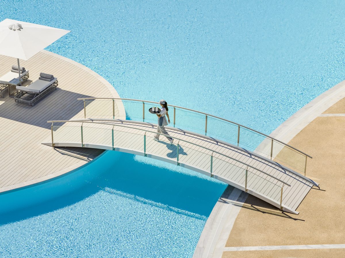 A member of staff carrying a tray across a slim footbridge that spans the pool, with a parasol and loungers on the deck.