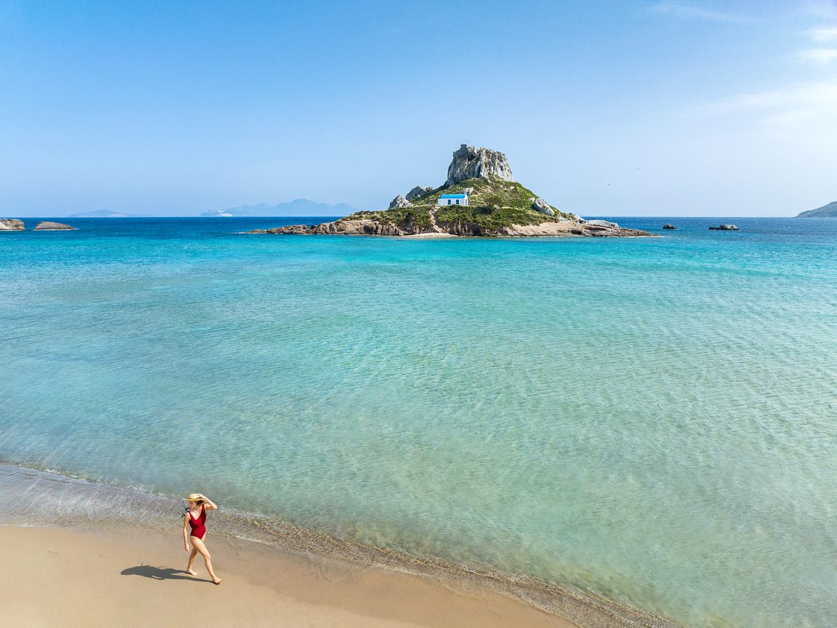 The pale sand of Agios Stefanos running along the shallow turquoise water, with the rocky Kastri islet offshore.