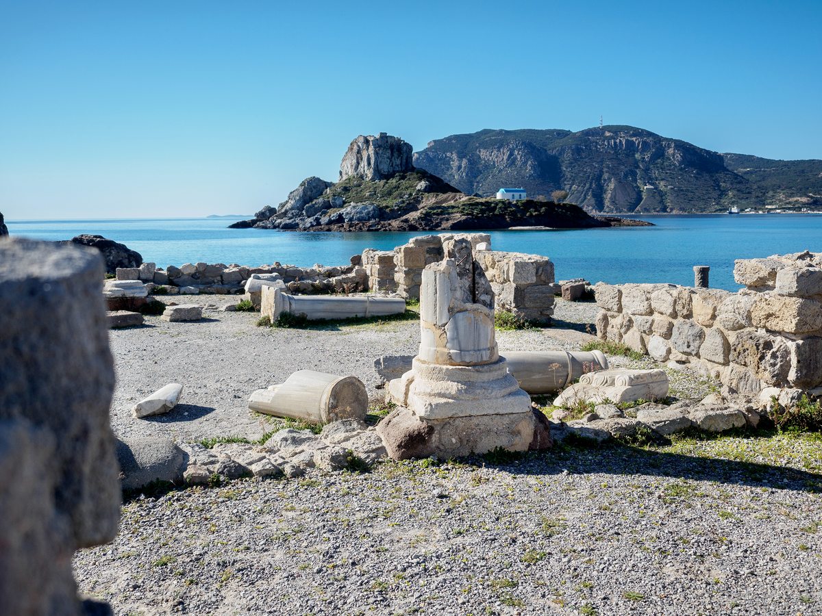 The low stone remains of an early-Christian basilica standing on the sand at the end of the beach, with the islet behind.