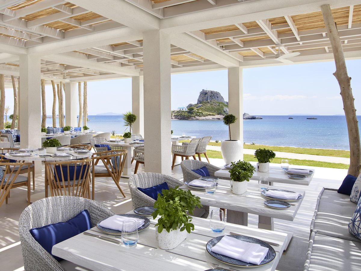 Ouzo set out under a white colonnade beside the sand, tables laid for dinner with the sea and the Kastri islet beyond.
