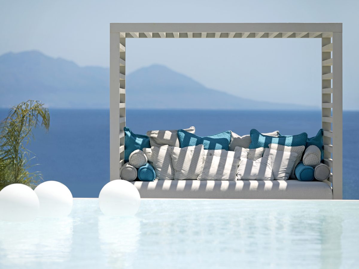 A white-framed daybed with striped and turquoise cushions on the pool deck, with the sea beyond.
