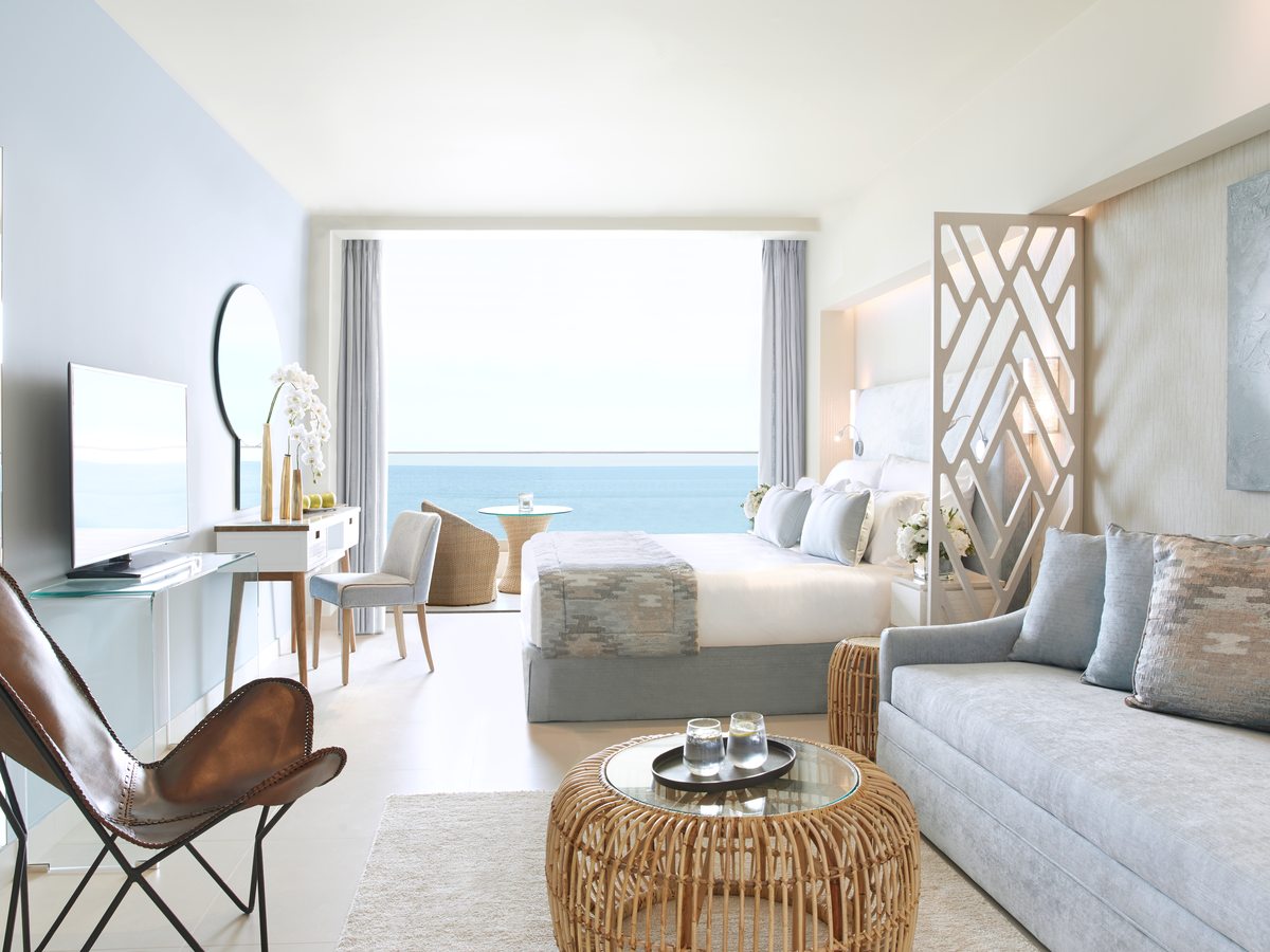 Open-plan suite with a bed, a sofa and rattan chairs, and glass doors open to a balcony above the sea.
