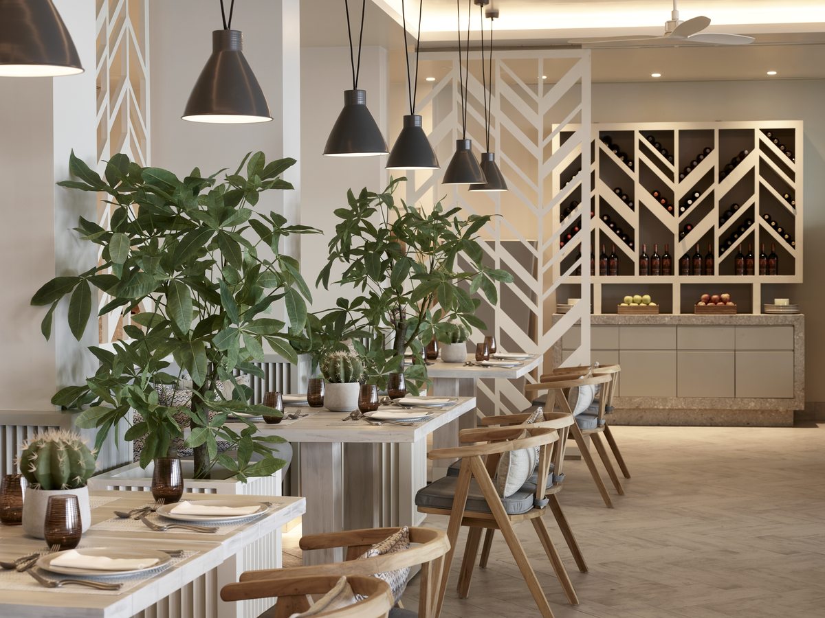 Dining room with laid tables, tall potted plants and pendant lights in front of a herringbone timber wall.