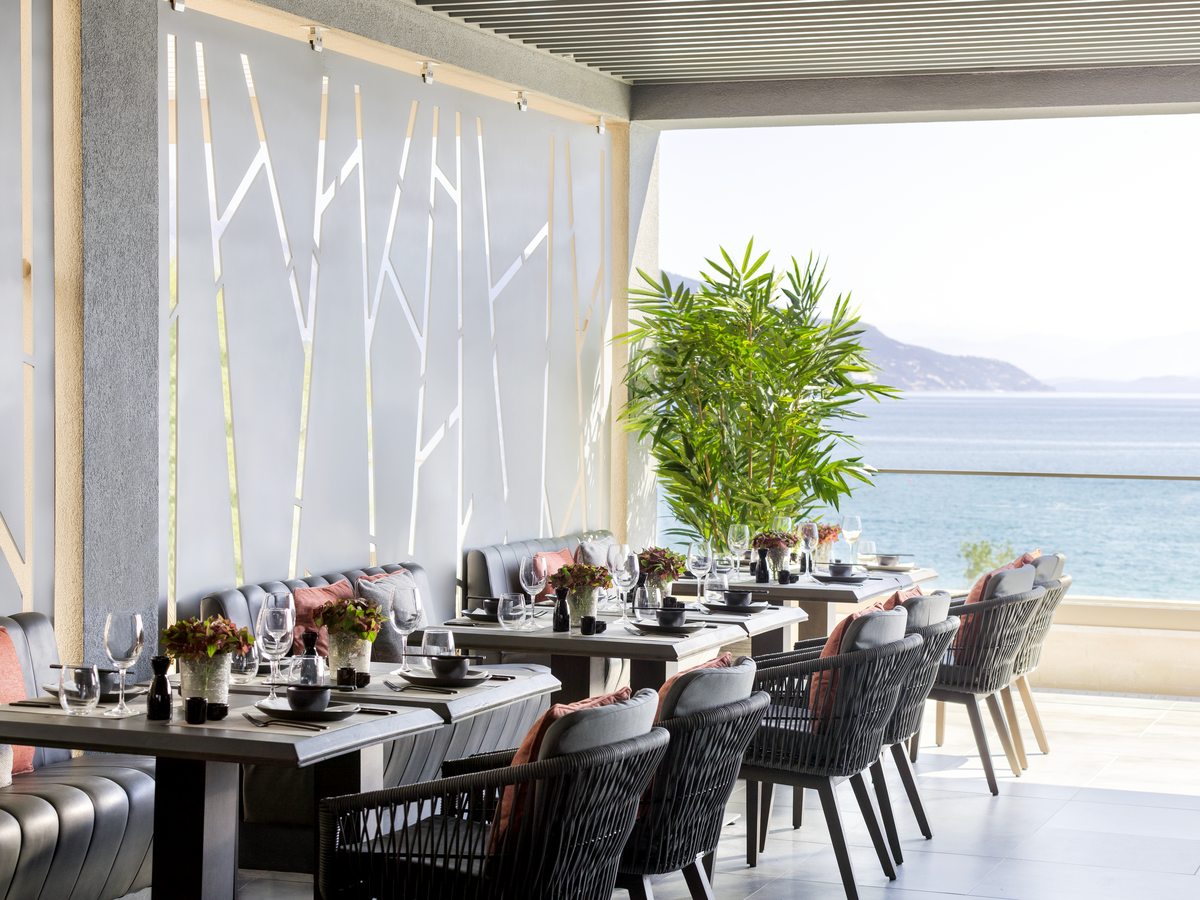 Terrace dining room with dark tables and chairs set for dinner, open to the hills and the sea beyond.