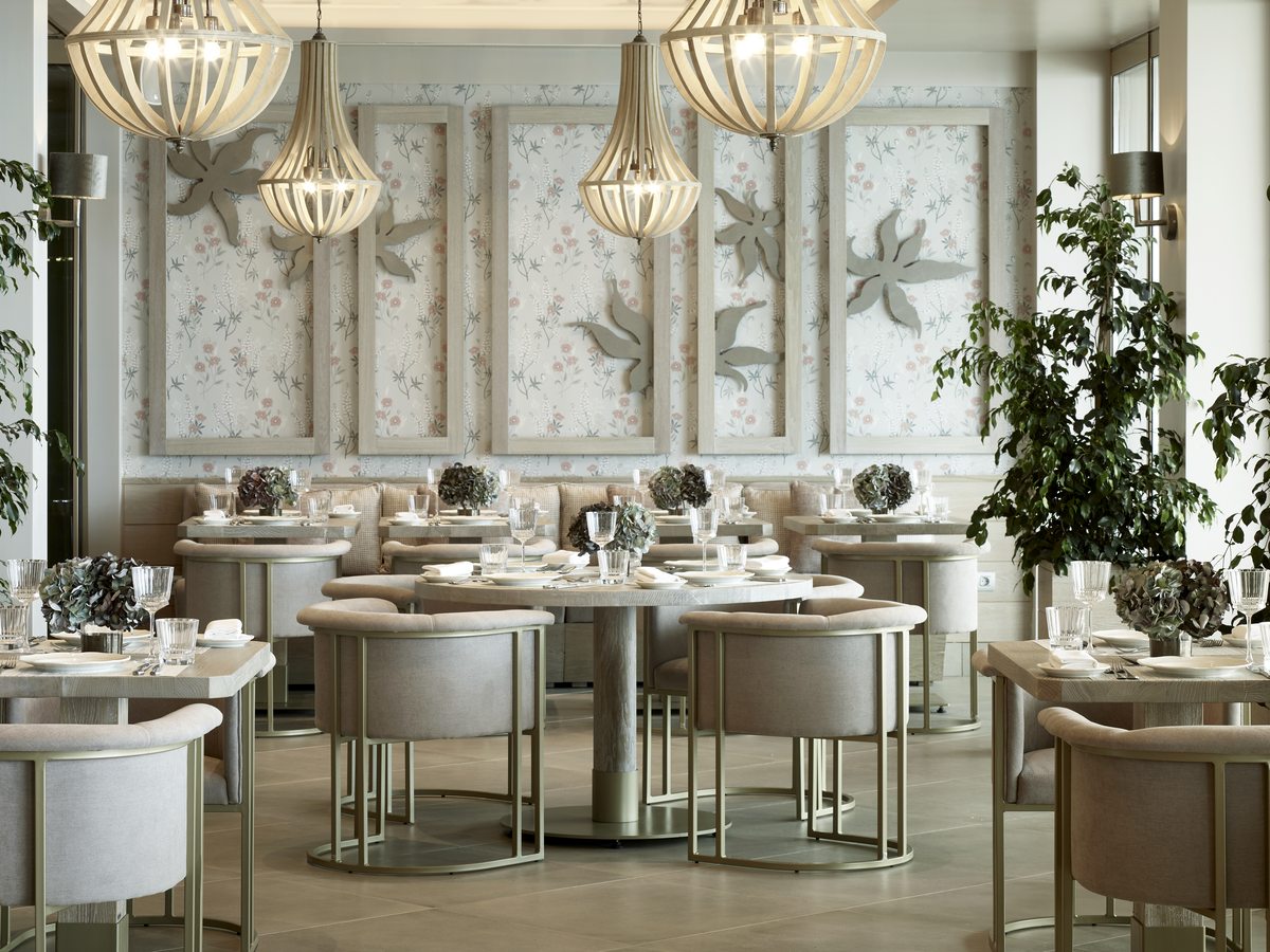 Formal dining room with round laid tables, cream chairs and chandeliers below a decorated panelled wall.