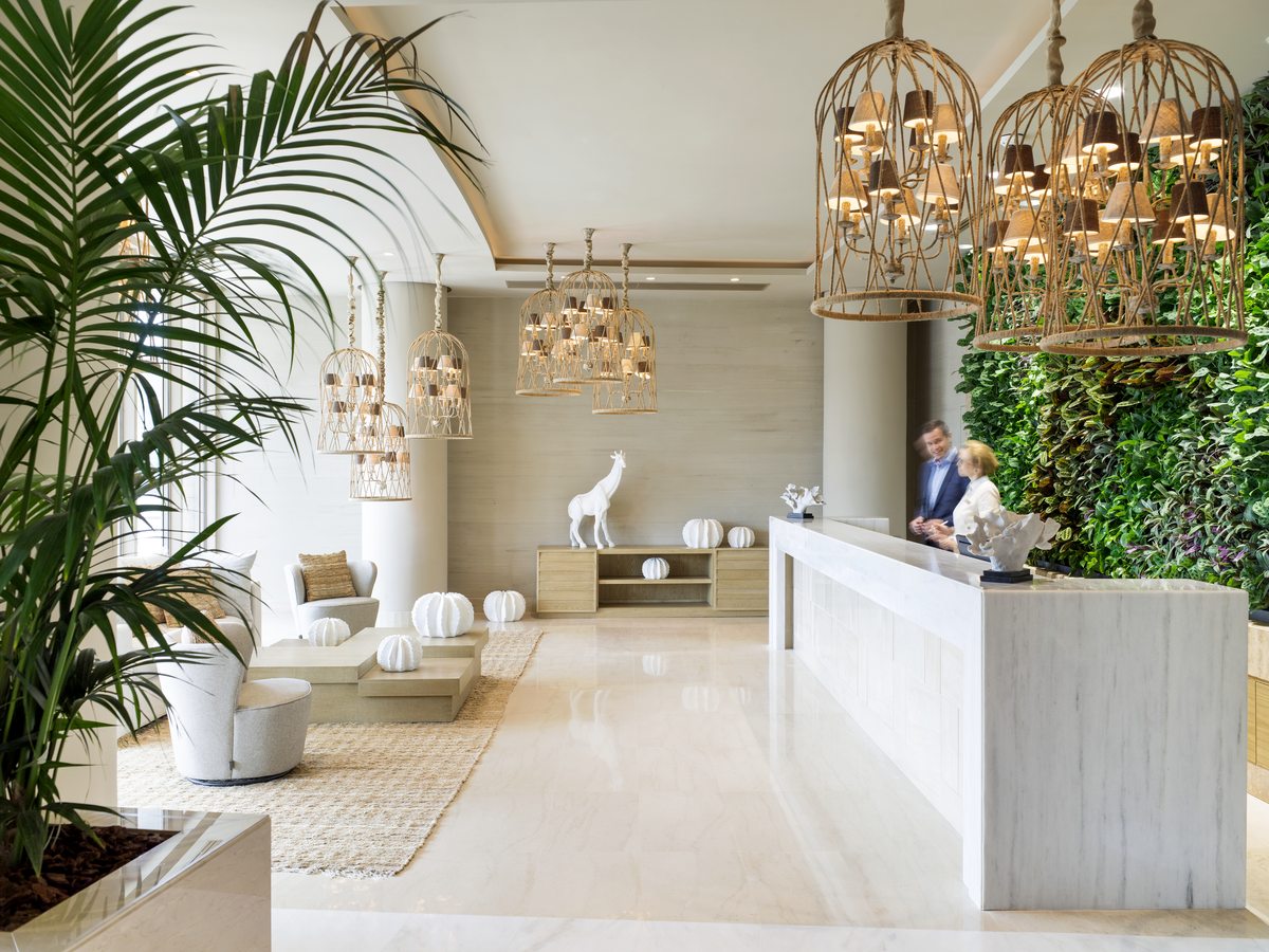Reception desk in a bright marble lobby, with cage pendant lights, a living green wall and tall palms.