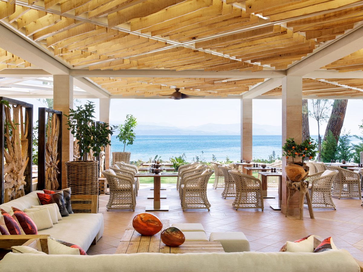 Open-sided dining room under a timber ceiling with ceiling fans, cushioned seating and tables facing the water.