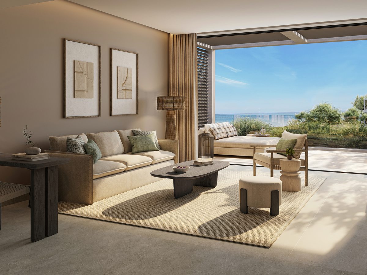 The lounge of a Deluxe One Bedroom Suite at Ikos Kissamos, with a long sofa, low tables, framed prints and doors onto a terrace with the sea beyond.