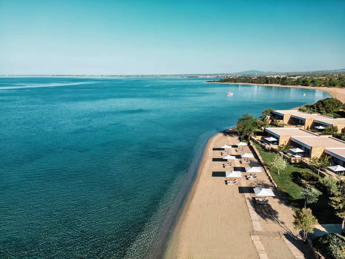 Rows of sunbeds and thatched parasols along the long sandy beach at Ikos Kissamos, with low room buildings and trees behind them.