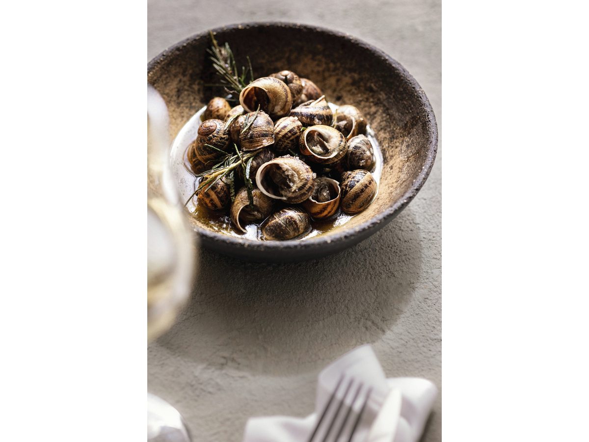 A dark bowl of cooked snails with sprigs of rosemary, set on a linen napkin beside a fork.