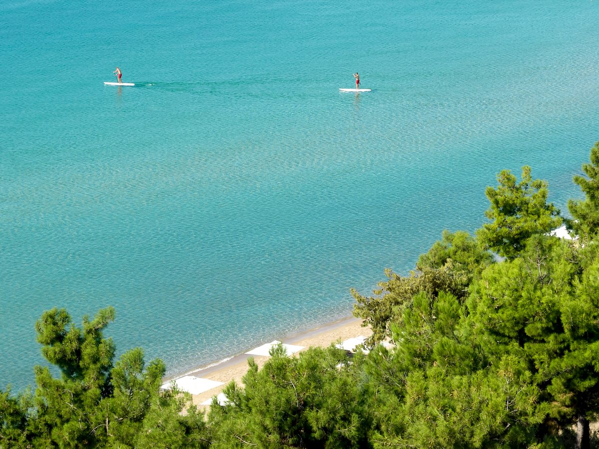 Two paddleboarders on flat turquoise water off the resort beach, with pines along the shore.
