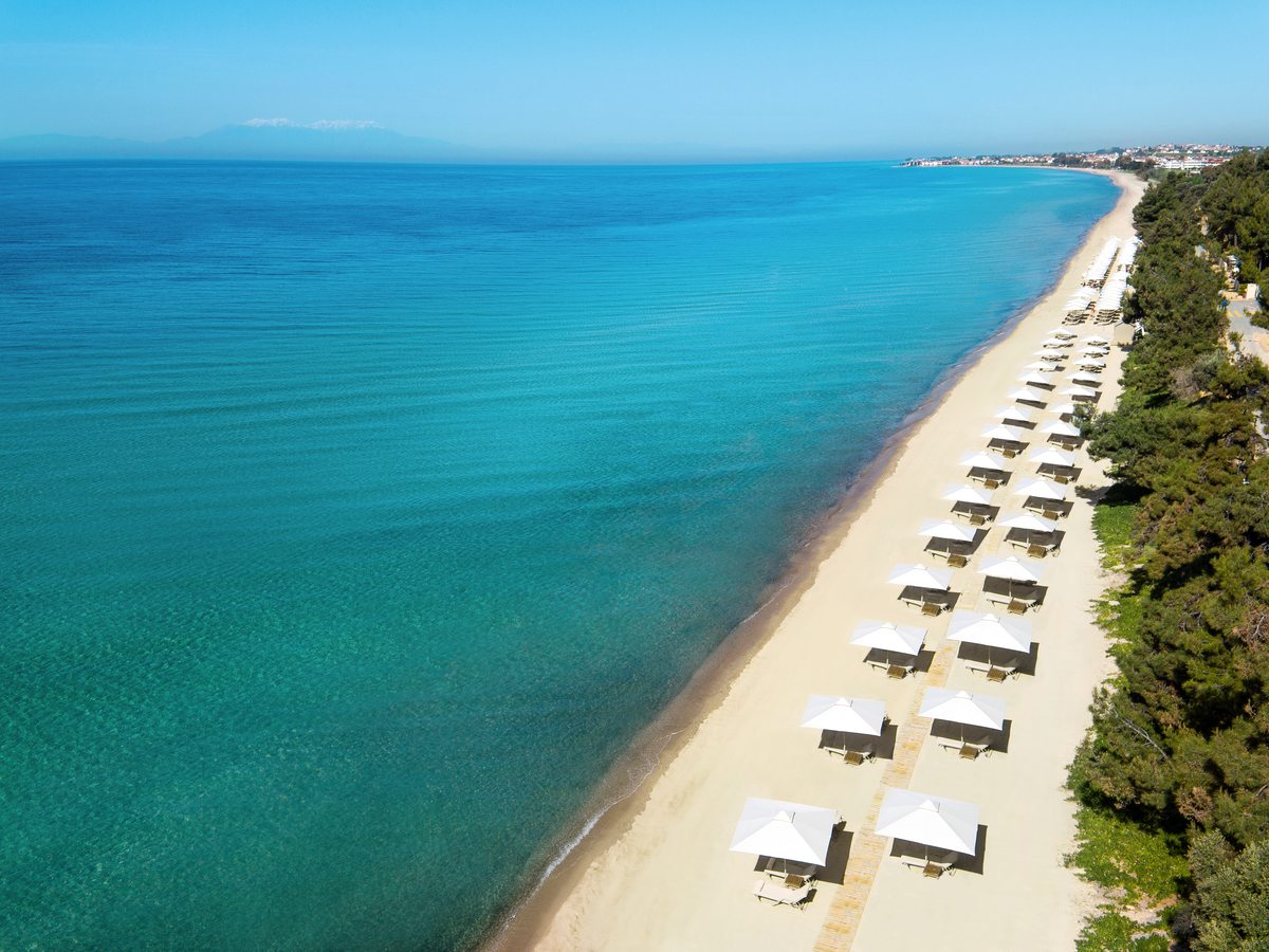 Aerial view of the resort beach, a long strip of pale sand lined with parasols and sunbeds beside clear turquoise shallows.