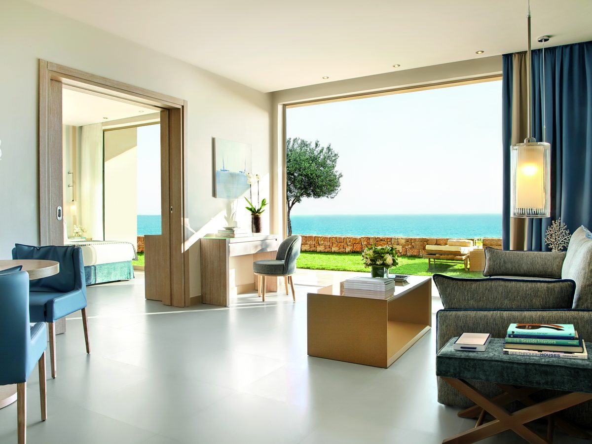 The living and dining area of a Deluxe Two Bedroom Family Suite, with sofas, a dining table and a bedroom door to one side, opening onto a lawn and the sea.