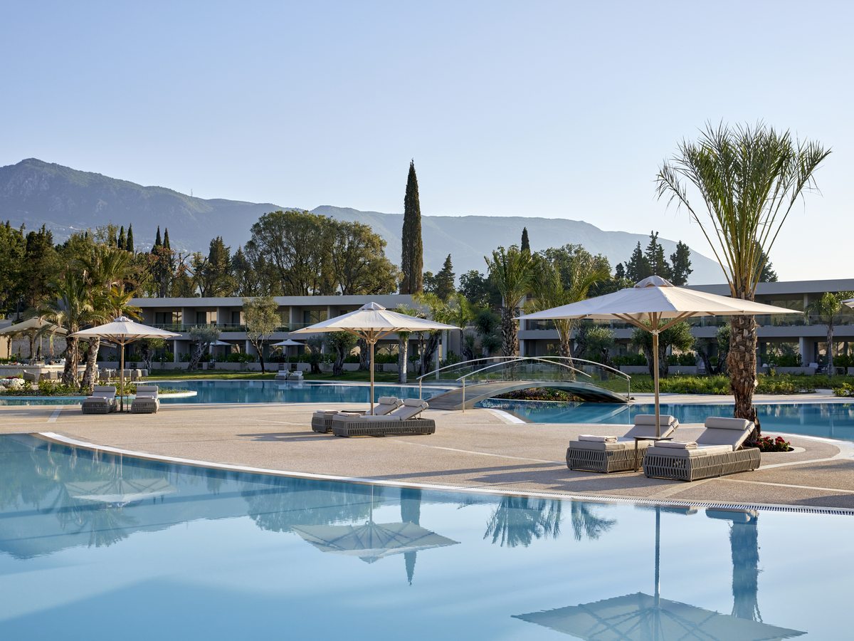 A curved pool with parasols, daybeds and palms around the edge, and wooded hills in the distance.