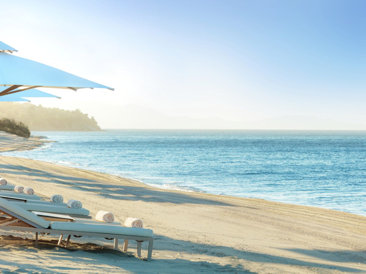 Sunbeds and a shade sail on the sand at Ikos Olivia, with the calm sea and the coastline beyond.