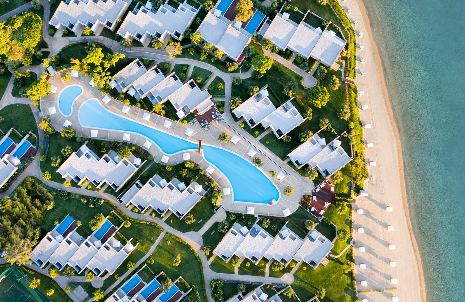 Ikos Olivia from the air: rows of single-storey bungalows spread flat around a lagoon pool right beside a sandy beach and the sea.