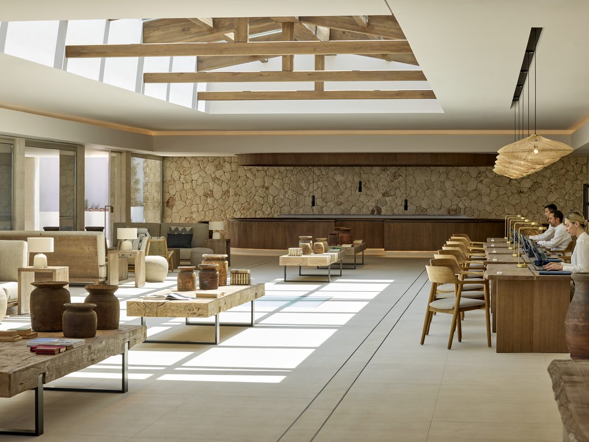 An open lobby lounge with pale seating, low tables and woven pendant lights under a beamed roof, and a reception desk along one side.