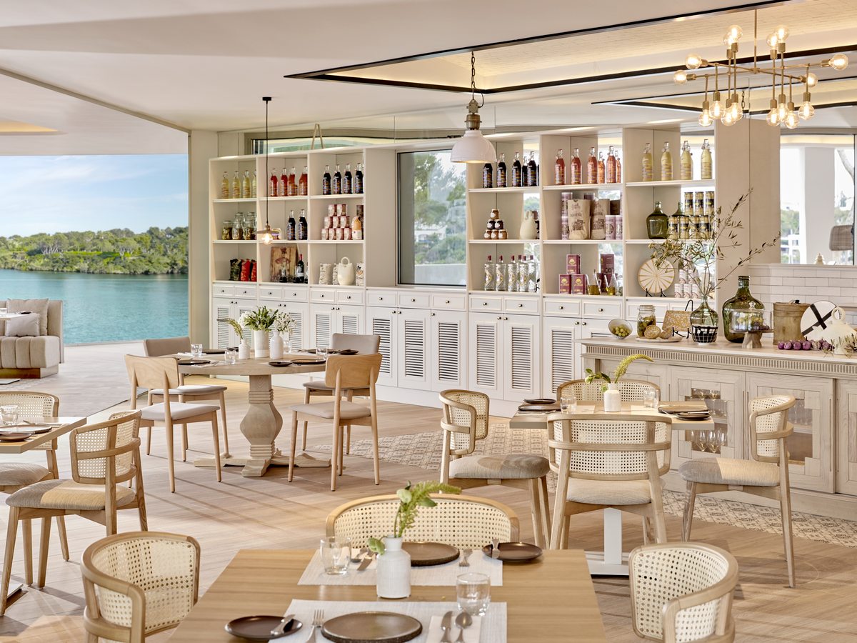 The dining room at Oliva, pale timber tables and cane chairs in front of a long bar of shelved bottles, with the cove through the windows.