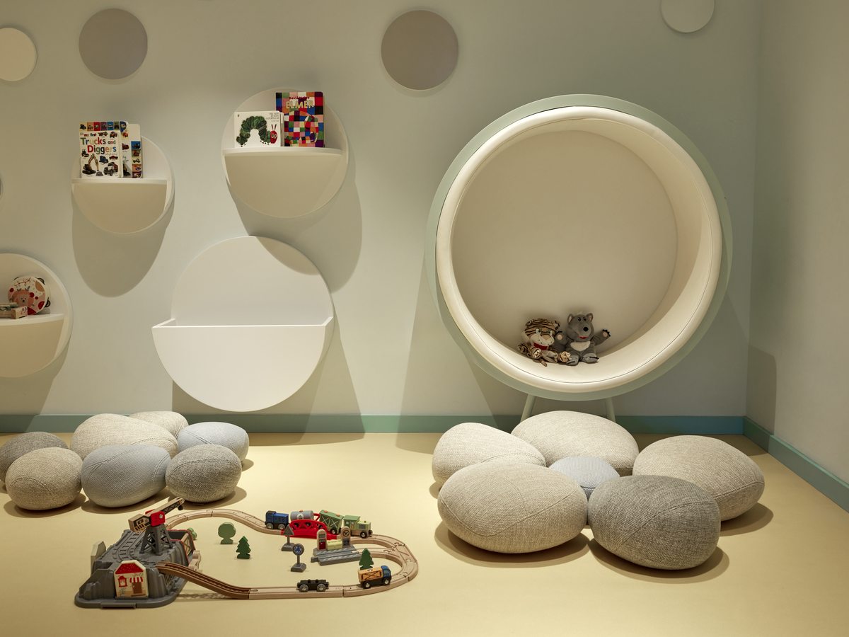 A play area in the baby centre with round wall alcoves holding books and toys, floor cushions and a wooden train set.